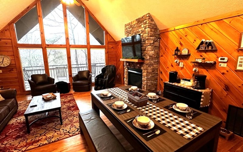 Relax in the family room at Lazy Bear Den, where a cozy stone fireplace and large windows frame peaceful wooded mountain views—the perfect spot to unwind and take in the scenery
